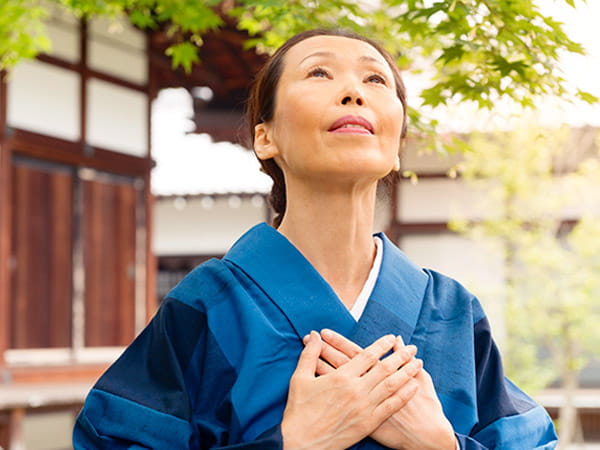 A mature Japanese woman wearing a blue kimono has her hands resting over her heart in a healing gesture as she looks up toward the sky. A mature Japanese woman wearing a blue kimono has her hands resting over her heart in a healing gesture as she looks up toward the sky.